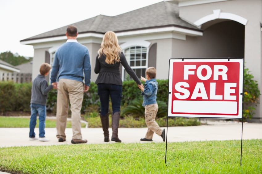 family holding hands looking at the fron of a house with for sale sign in front